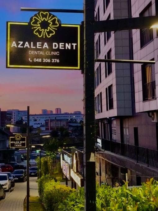 Tabela e ndriçuar e Azalea Dent mbi rrugë në mbrëmje, me logon e azaleas, emrin e klinikës dhe numrin e telefonit, para qiellit në perëndim.